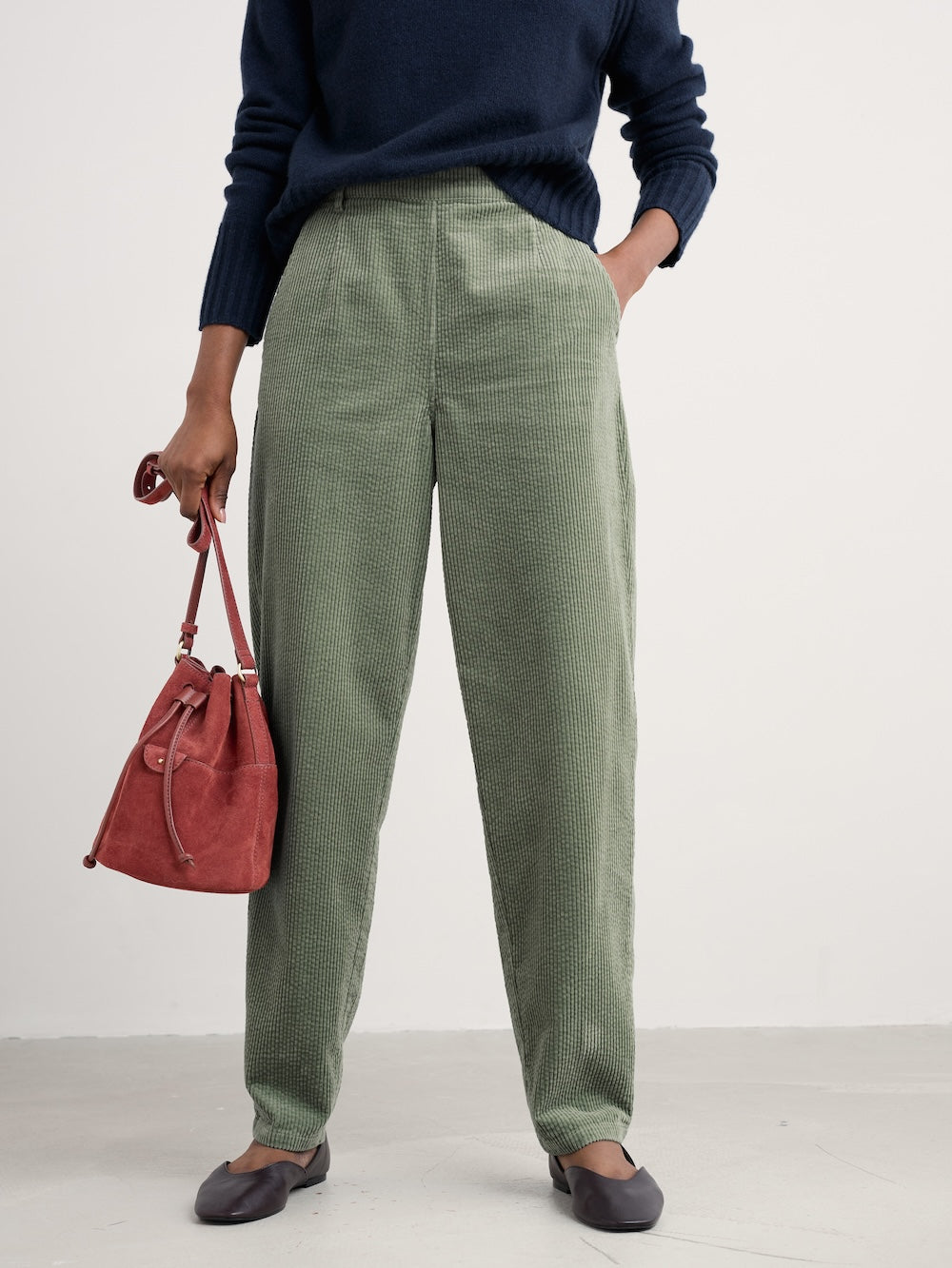 Person wearing green pants and a navy sweater, holding a red bag on a white background