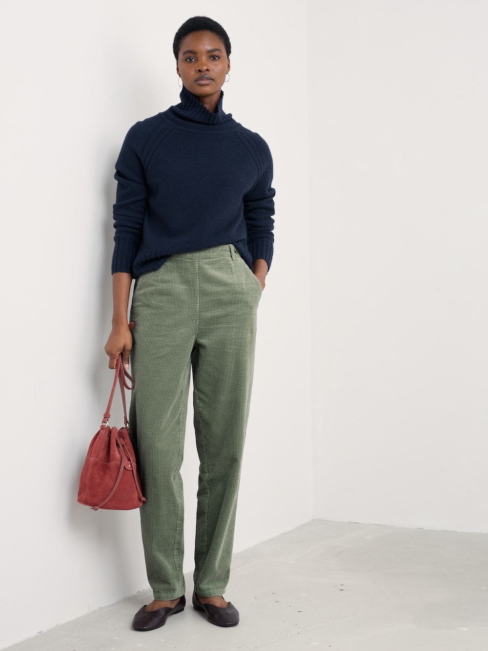 Person wearing a navy sweater and green pants, holding a red bag against a white background