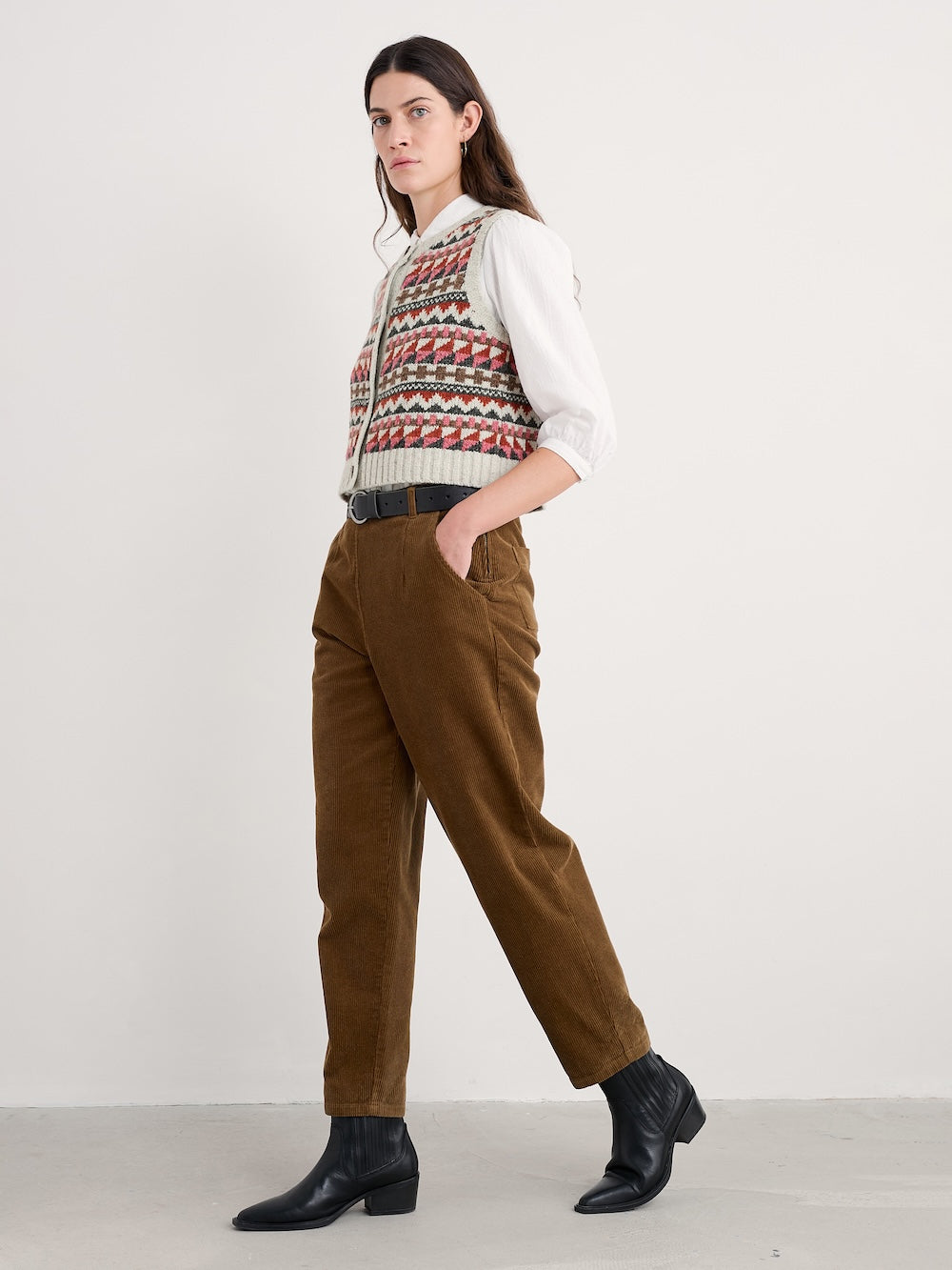 Woman wearing a patterned vest, white shirt, brown pants, and black shoes on a white background