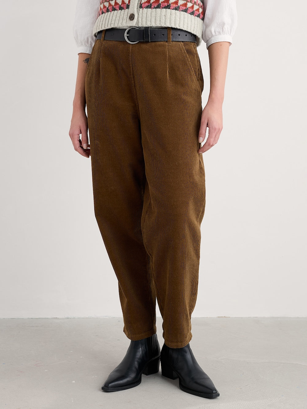 Person wearing brown corduroy pants with a white sweater and black boots on a plain background