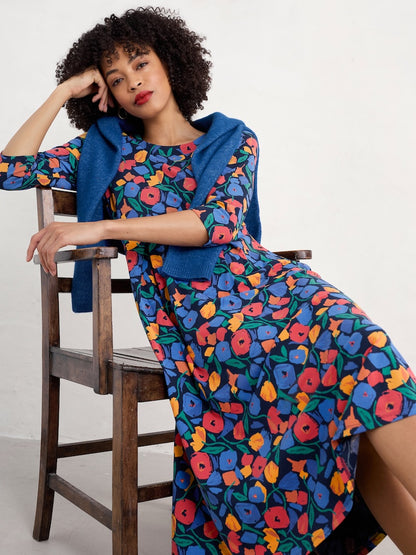 Woman wearing a colourful floral dress with a blue cardigan sitting on a wooden chair.