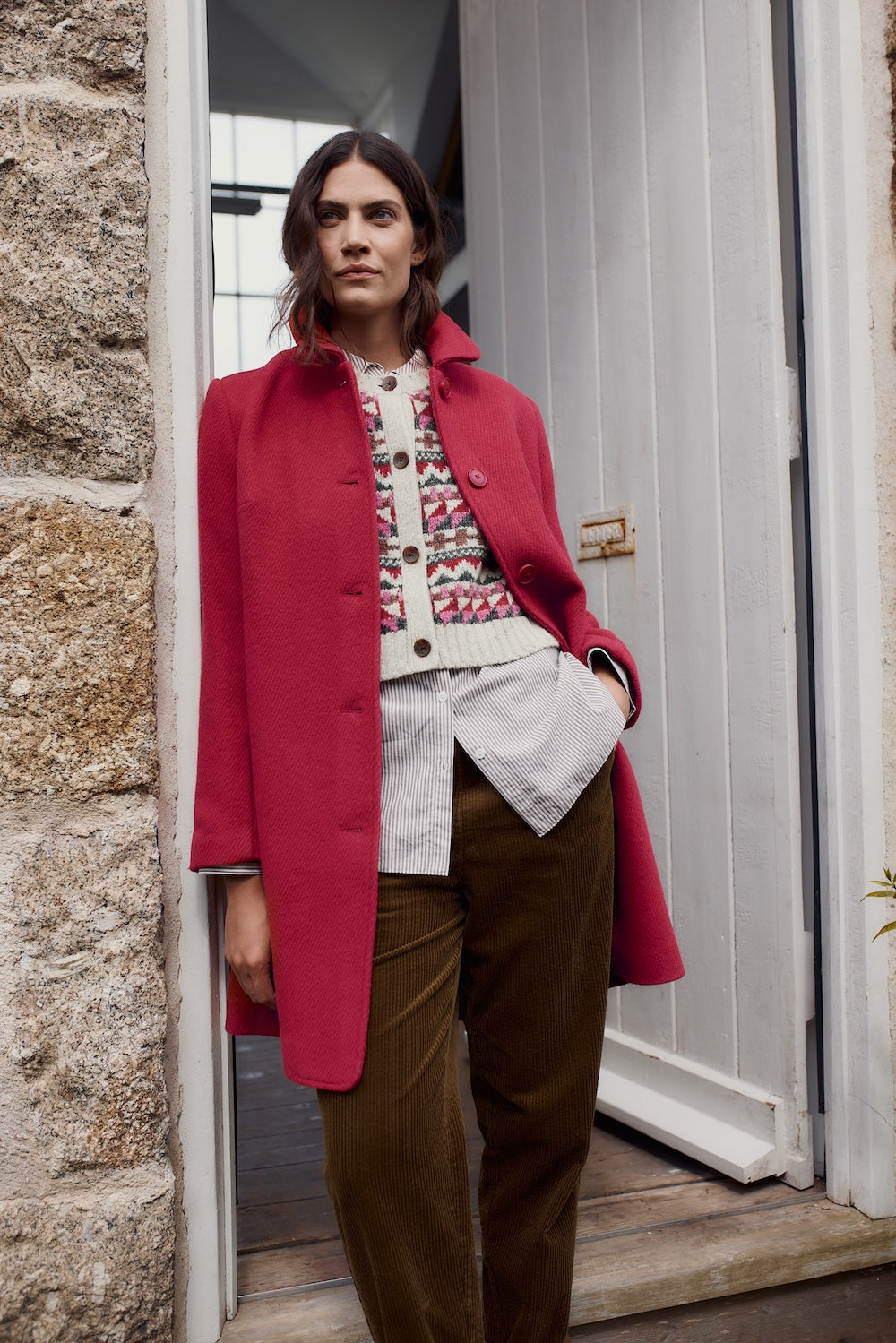 Person wearing a pink coat standing in front of a stone wall and white door.