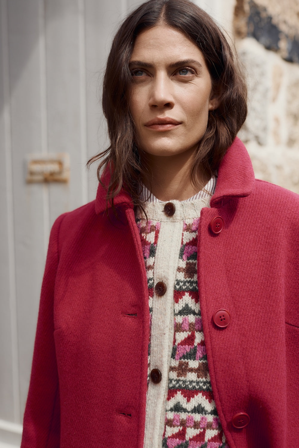 Woman wearing a pink coat over a patterned sweater, standing against a neutral background.