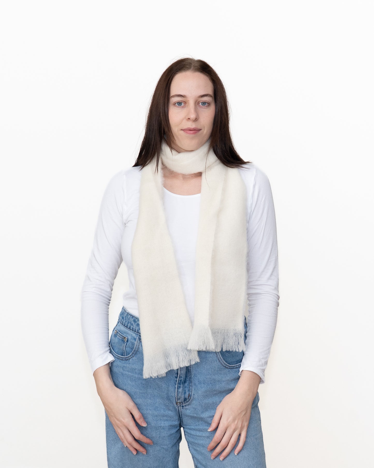 A woman modeling a cream-colored, fringed faux fur scarf over a white long-sleeve top and blue jeans, shot against a plain white studio background.