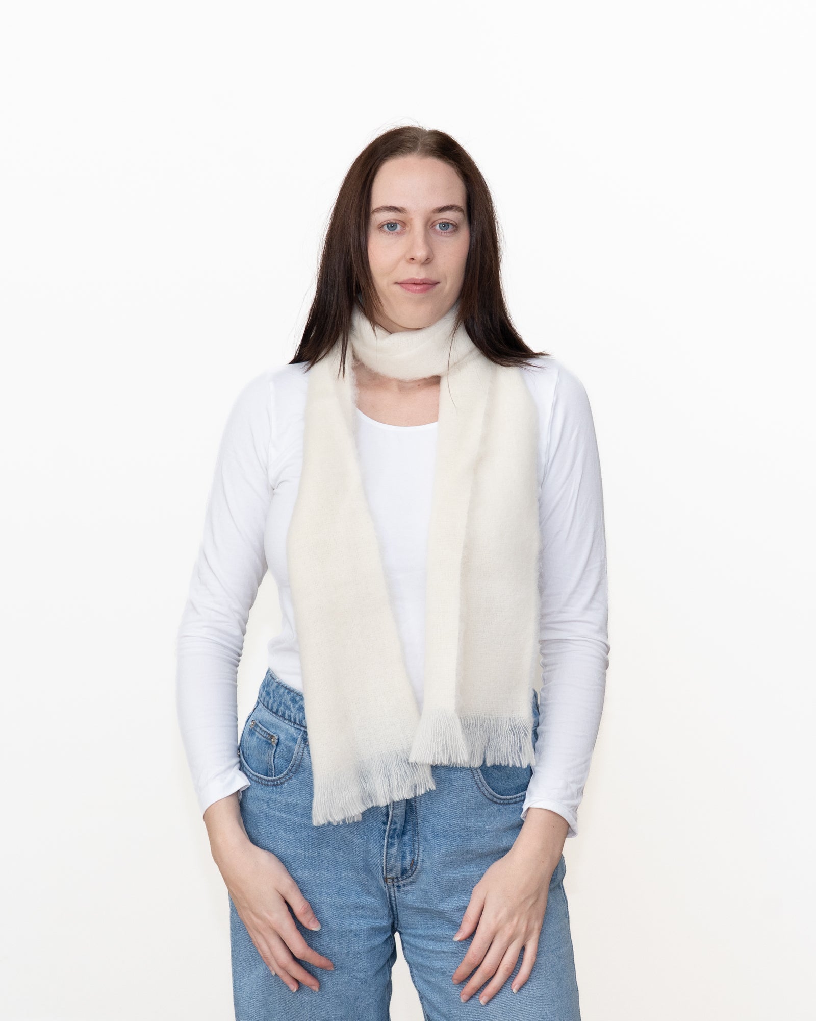 A woman modeling a cream-colored, fringed faux fur scarf over a white long-sleeve top and blue jeans, shot against a plain white studio background.