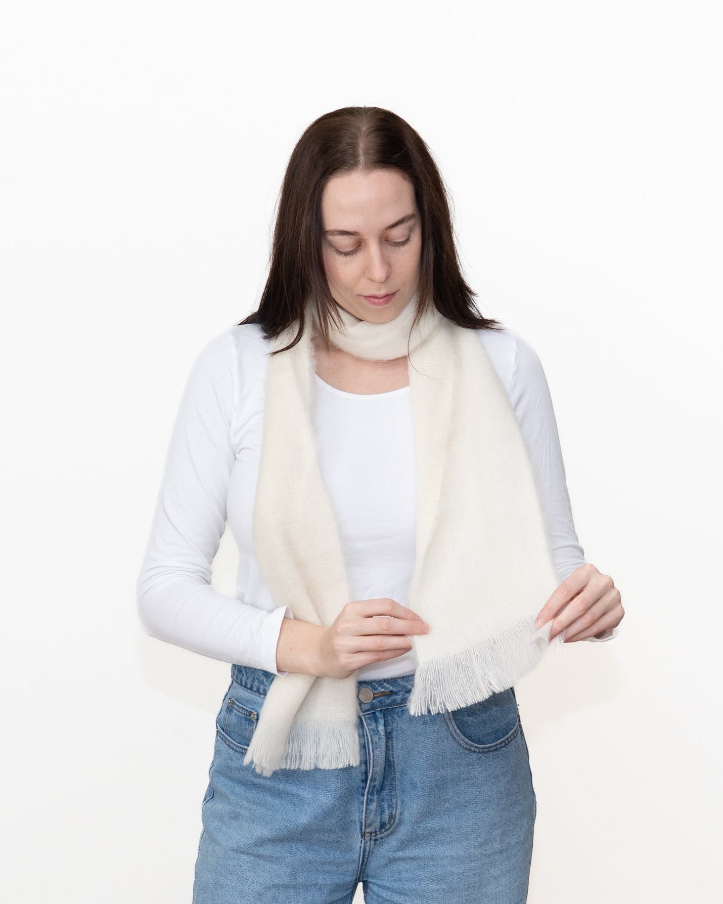 A woman wearing a cream-colored, soft-textured alpaca scarf with fringe, styled over a white long-sleeve top and blue jeans, against a plain white background.