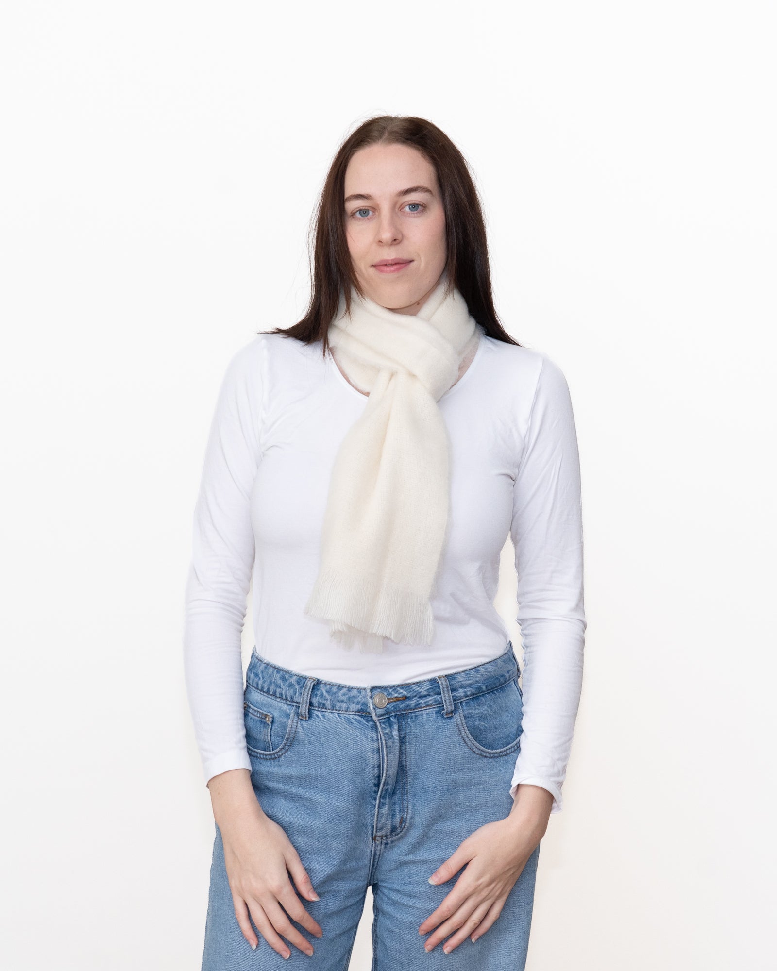 A woman modeling a soft cream-colored knit scarf wrapped around her neck, paired with a white long-sleeve top and blue jeans, against a plain white background.