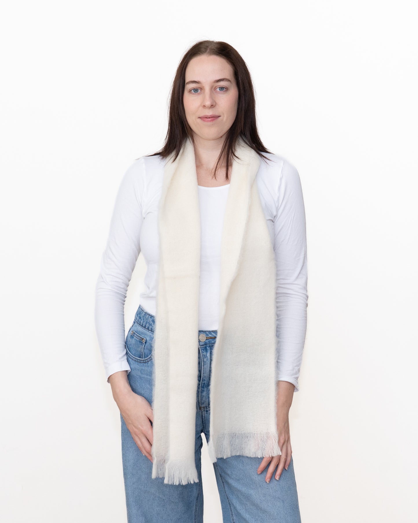 A woman modeling a long, cream-colored knit scarf with fringed ends, worn draped around her neck over a white long-sleeve top and blue jeans, against a plain white studio background.