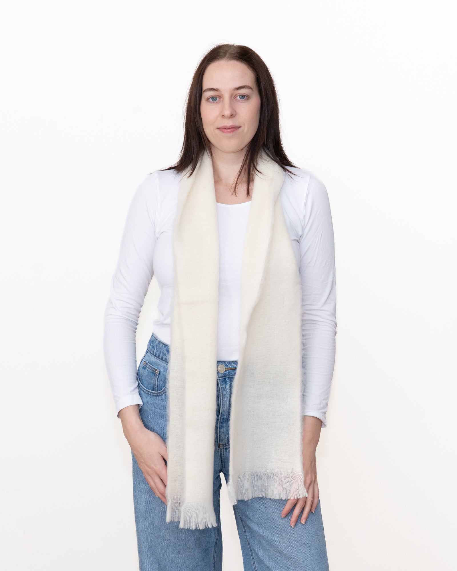 A woman modeling a long, cream-colored knit scarf with fringed ends, worn draped around her neck over a white long-sleeve top and blue jeans, against a plain white studio background.