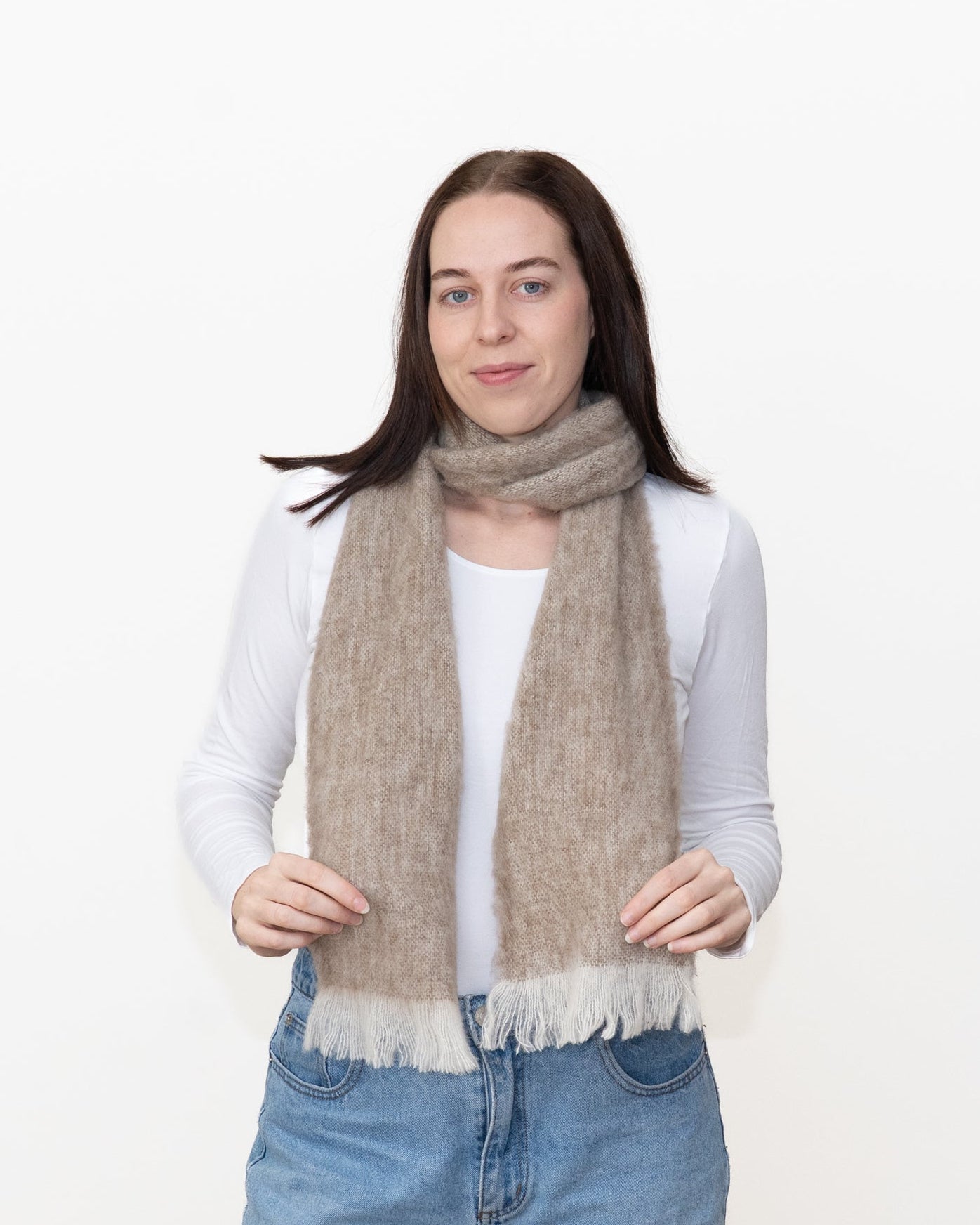 A woman modeling a soft, fuzzy knit scarf in a light taupe-beige color with white fringe, worn over a white long-sleeve top and blue jeans against a plain white background.