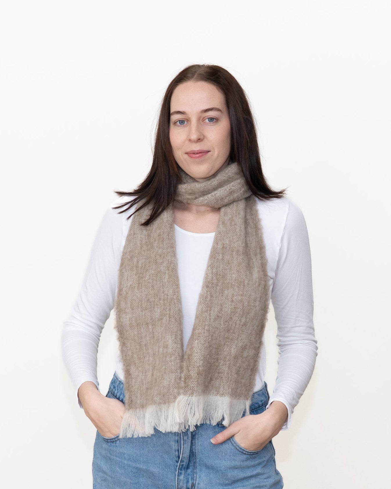 A woman wearing a light beige textured knit scarf with white fringe, styled over a white long-sleeve top and blue jeans, standing against a plain white background.