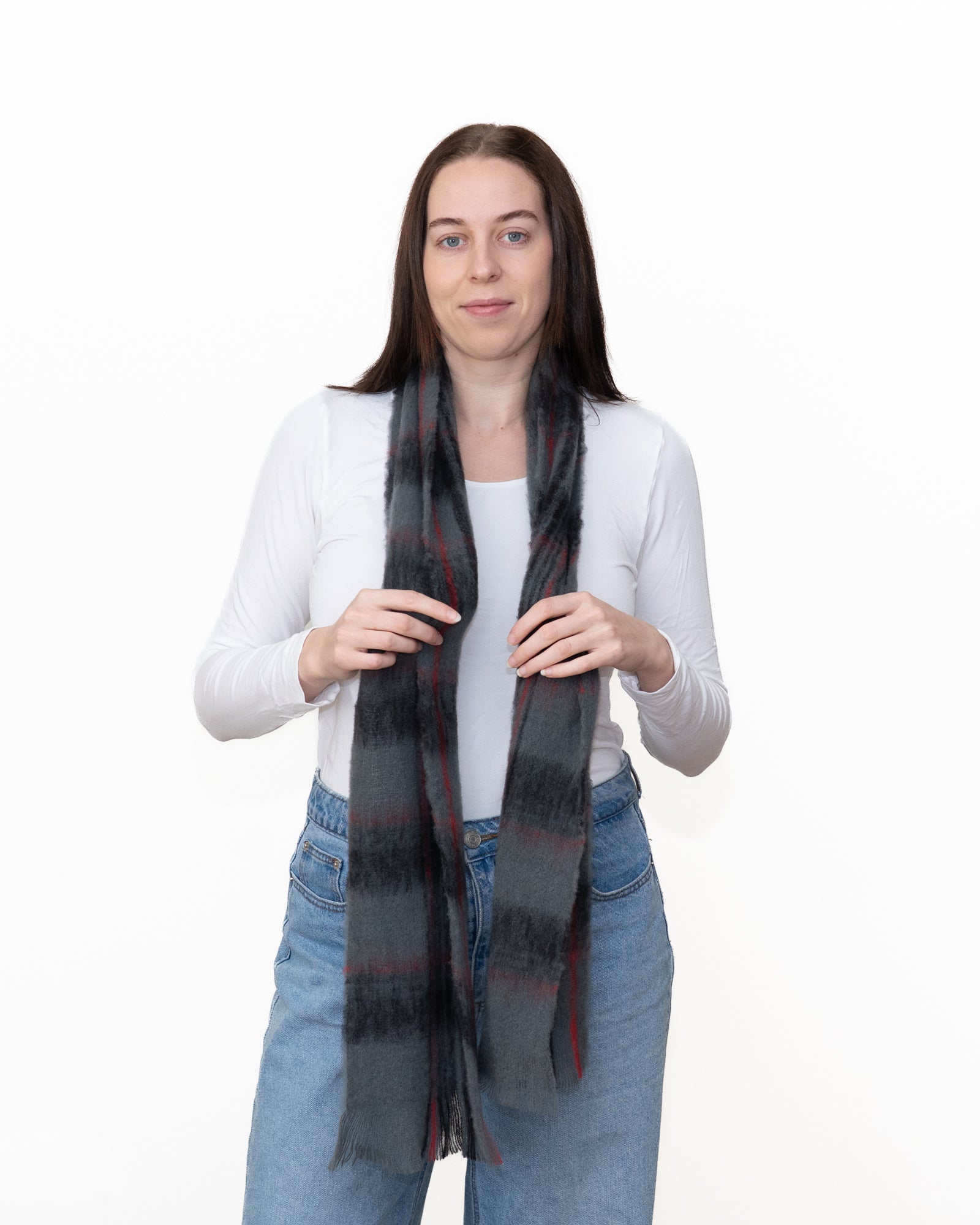 A woman modeling a dark gray and black plaid scarf with subtle red stripes, worn around her neck over a white long-sleeve top and blue jeans against a plain white background.