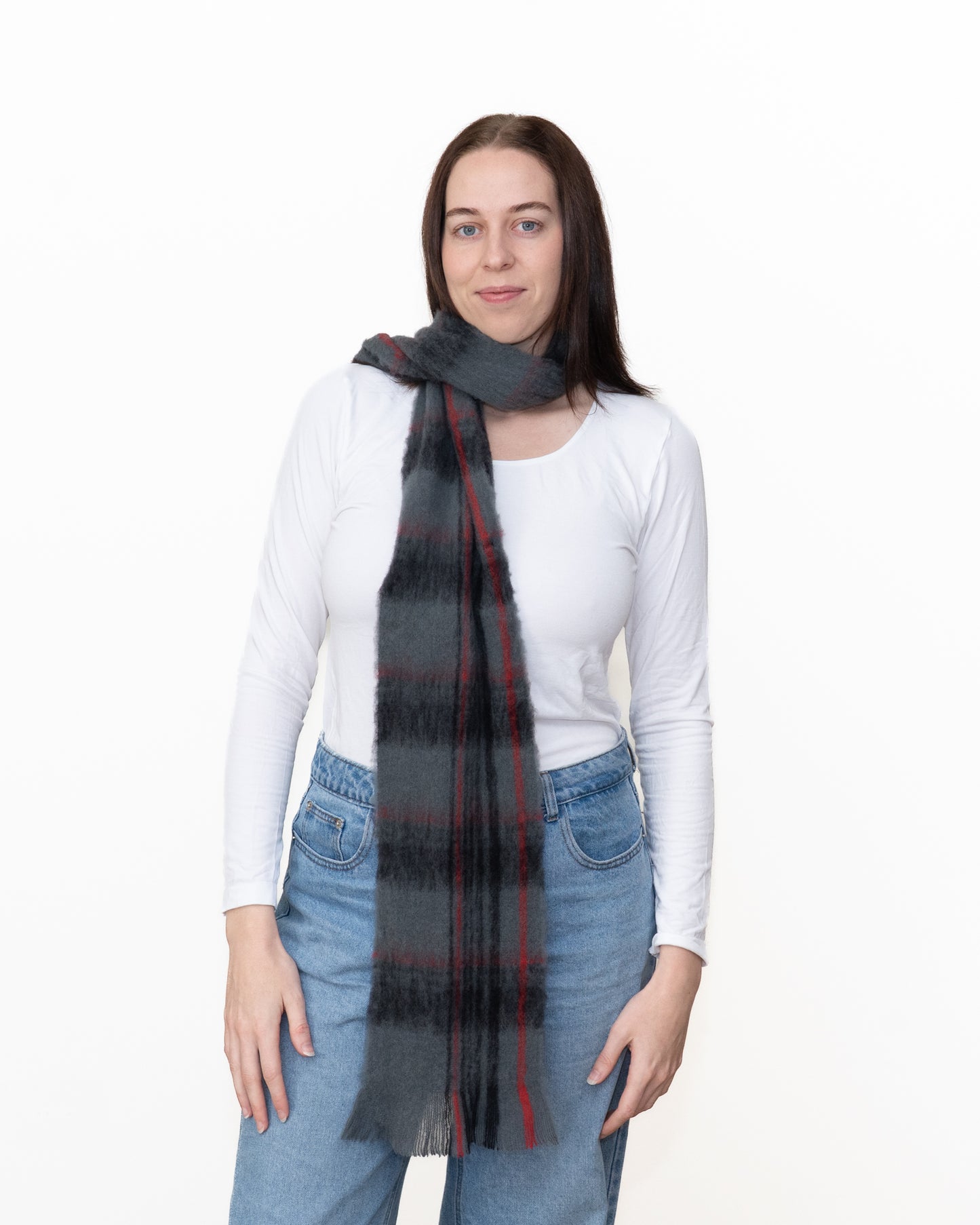 A woman modeling a dark gray and black plaid scarf with red stripes, worn around her neck over a white long-sleeve top and blue jeans against a plain white background.