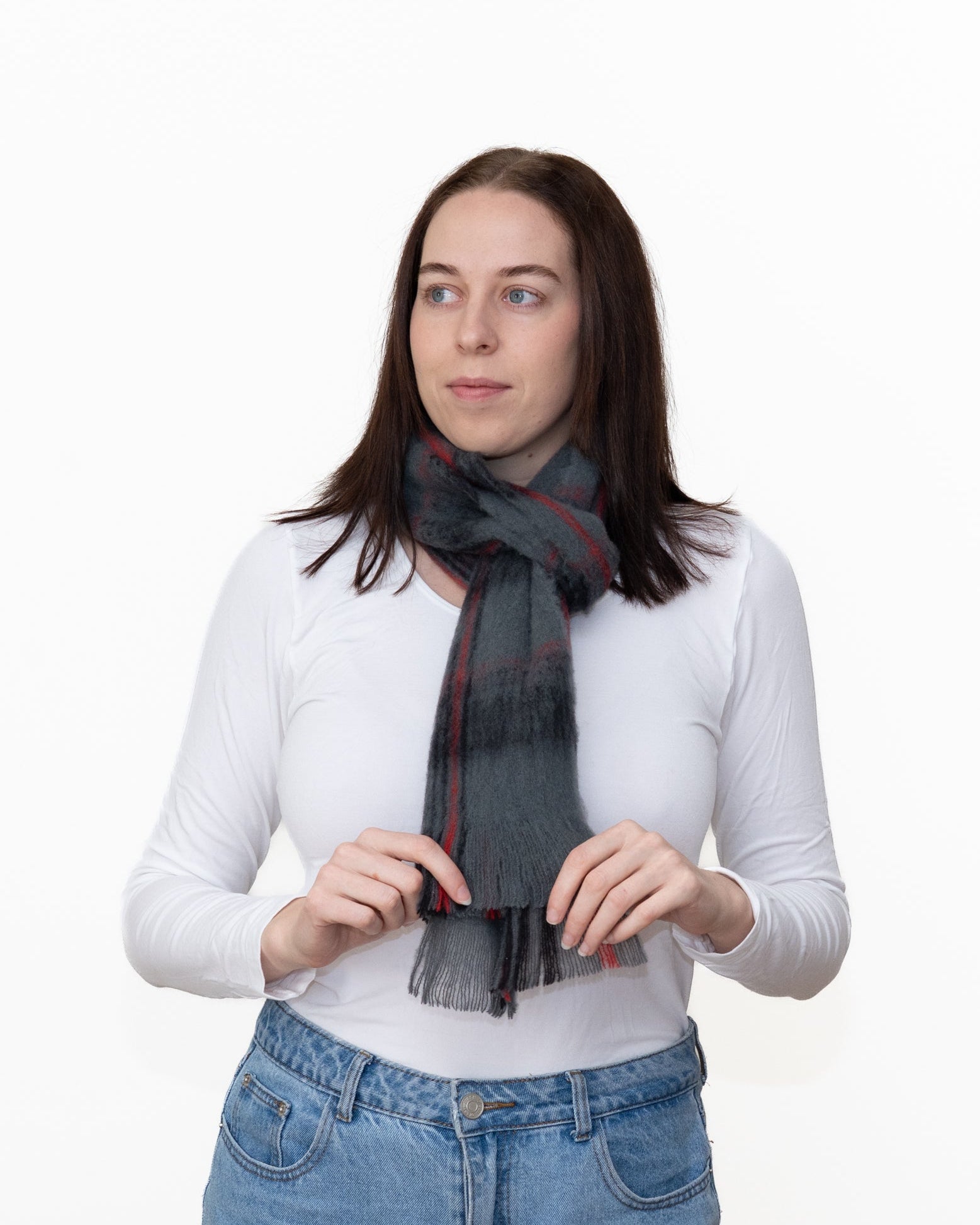 A woman with long brown hair wearing a white long-sleeve top and blue jeans, modeling a dark gray plaid scarf with red and black stripes against a plain white background.