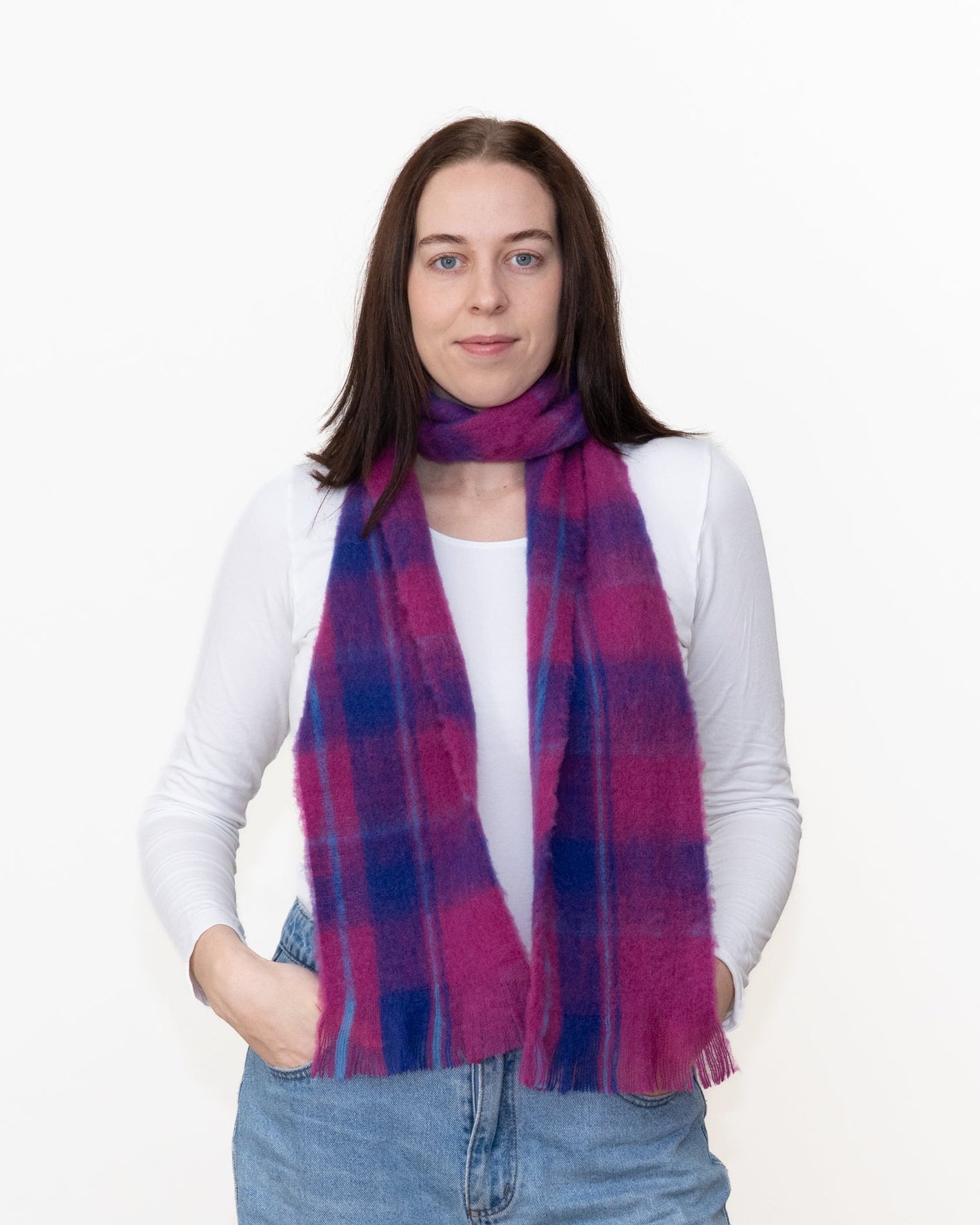 A woman wearing a soft, fuzzy plaid scarf in vibrant magenta pink and navy blue, styled over a white long-sleeve top and blue jeans against a plain white background.