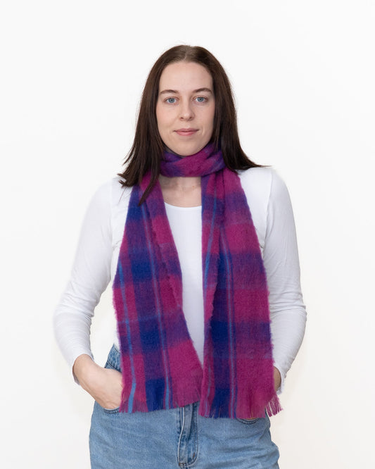 A woman wearing a soft, fuzzy plaid scarf in vibrant magenta pink and navy blue, styled over a white long-sleeve top and blue jeans against a plain white background.