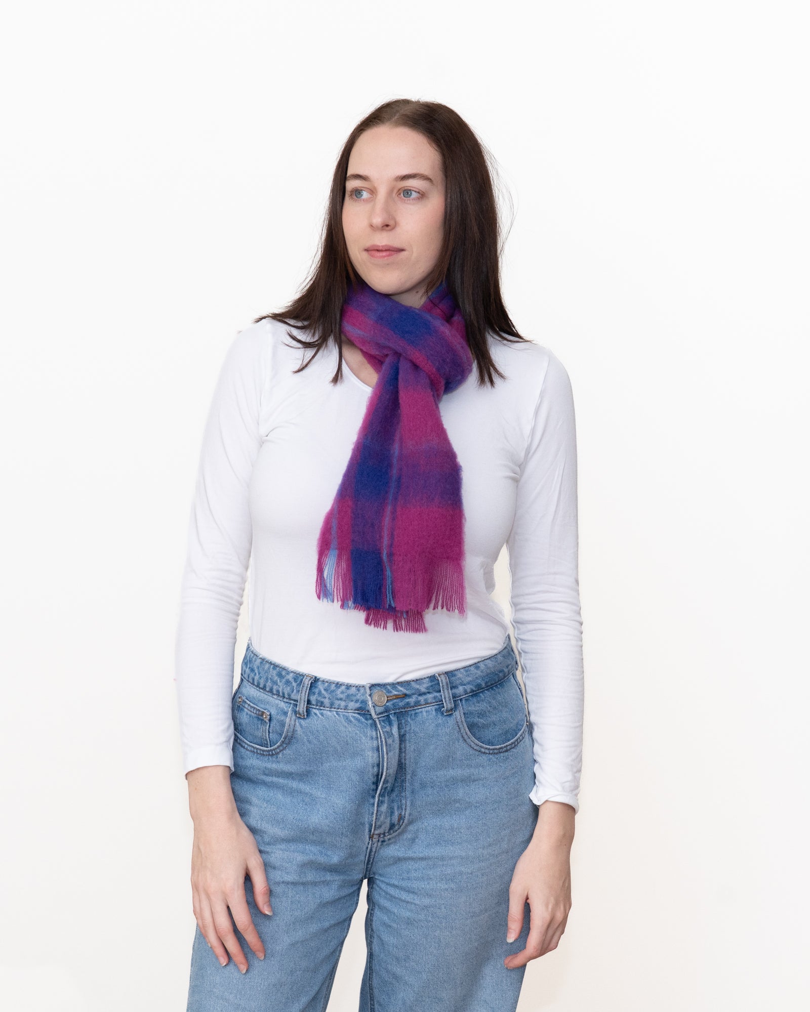 A woman wearing a white long-sleeve top and blue jeans, wrapped in a soft plaid scarf with purple, blue, and pink tones, against a plain white background.