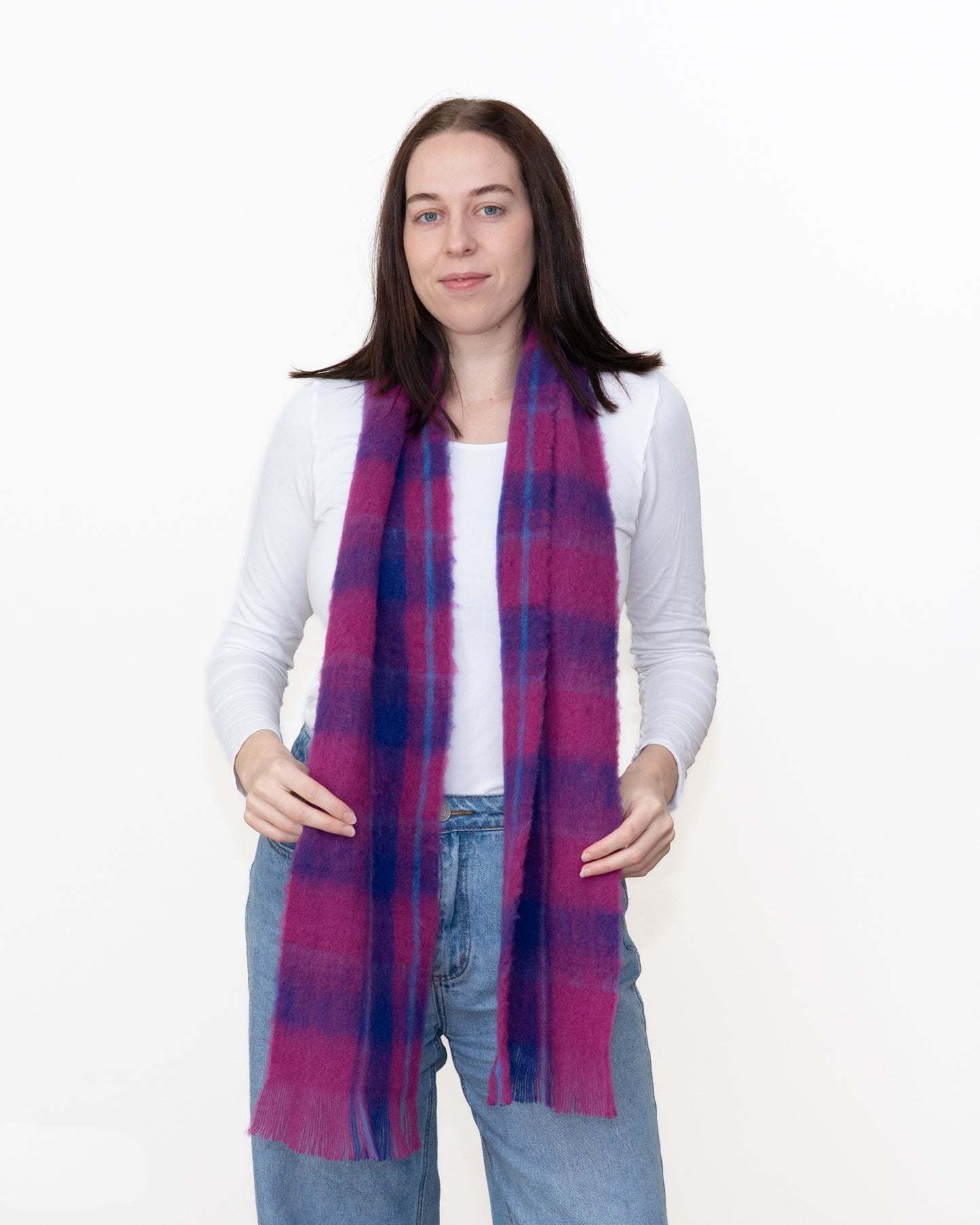 A woman modeling a lightweight plaid scarf in shades of magenta pink and dark blue, worn over a white long-sleeve top and blue jeans against a plain white background.