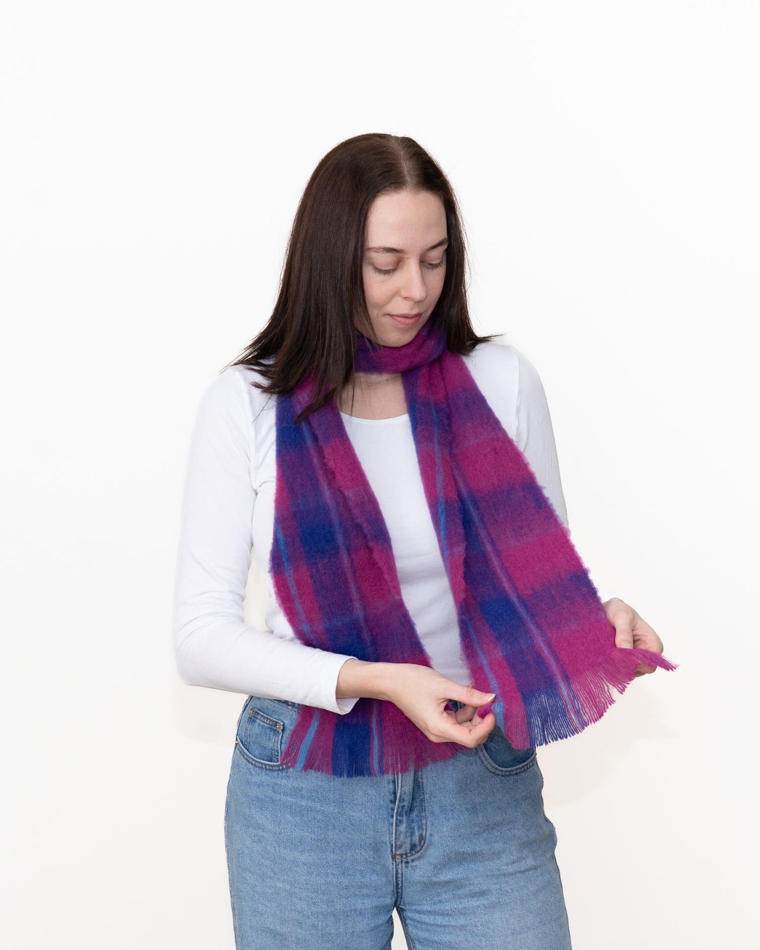 A woman wearing a white long-sleeve top and blue jeans models a fringed plaid scarf in purple, pink, and blue tones against a plain white background.