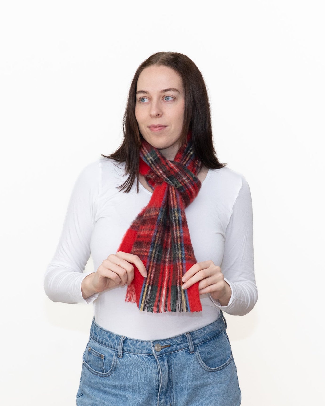 A woman wearing a red and black plaid scarf with green and yellow stripes, styled over a white long-sleeve top and blue jeans, standing against a plain white background.
