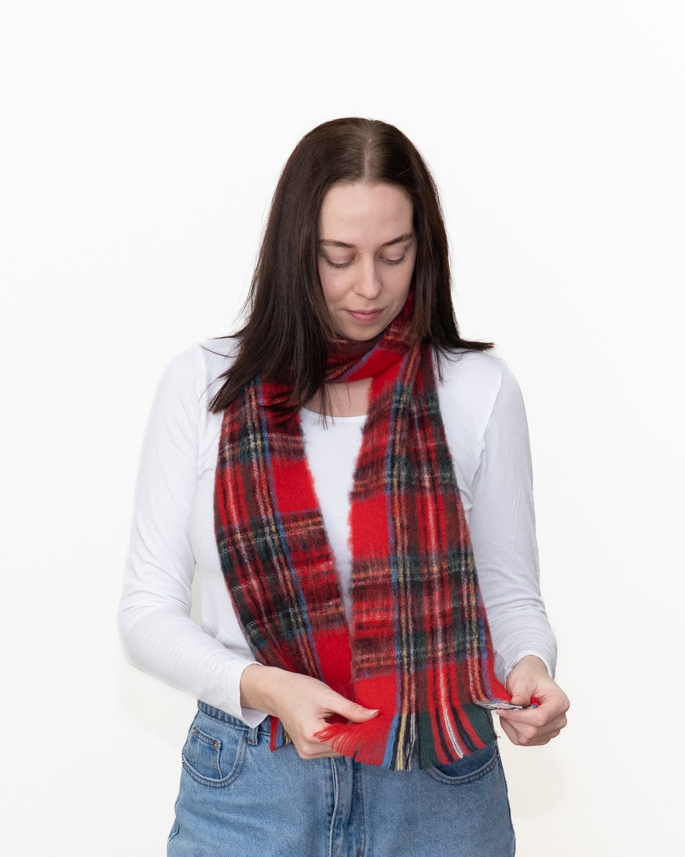 A woman wearing a red and black plaid scarf with blue jeans and a white long-sleeve top, standing against a plain white background.