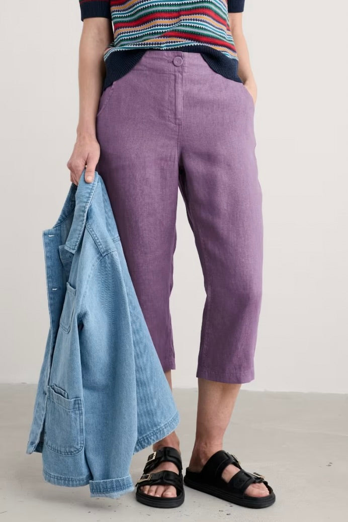 Woman wearing purple pants and a striped shirt, holding a denim jacket against a plain background