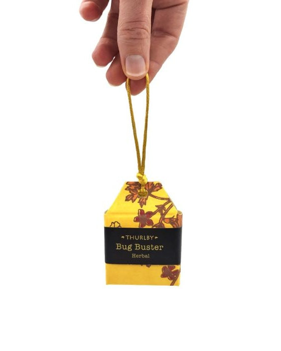 A hand holding a small yellow tea bag-shaped Bug Buster herbal air freshener by Thurlcy, featuring a black label and floral pattern, on a white background.