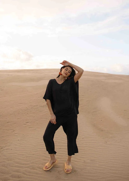 Woman wearing black bamboo pants and top standing on a sand dune with a neutral sky.