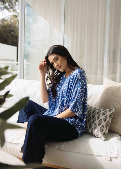 Woman sitting on a couch wearing a blue pattern shirt and navy blue bamboo pants, with a neutral background.
