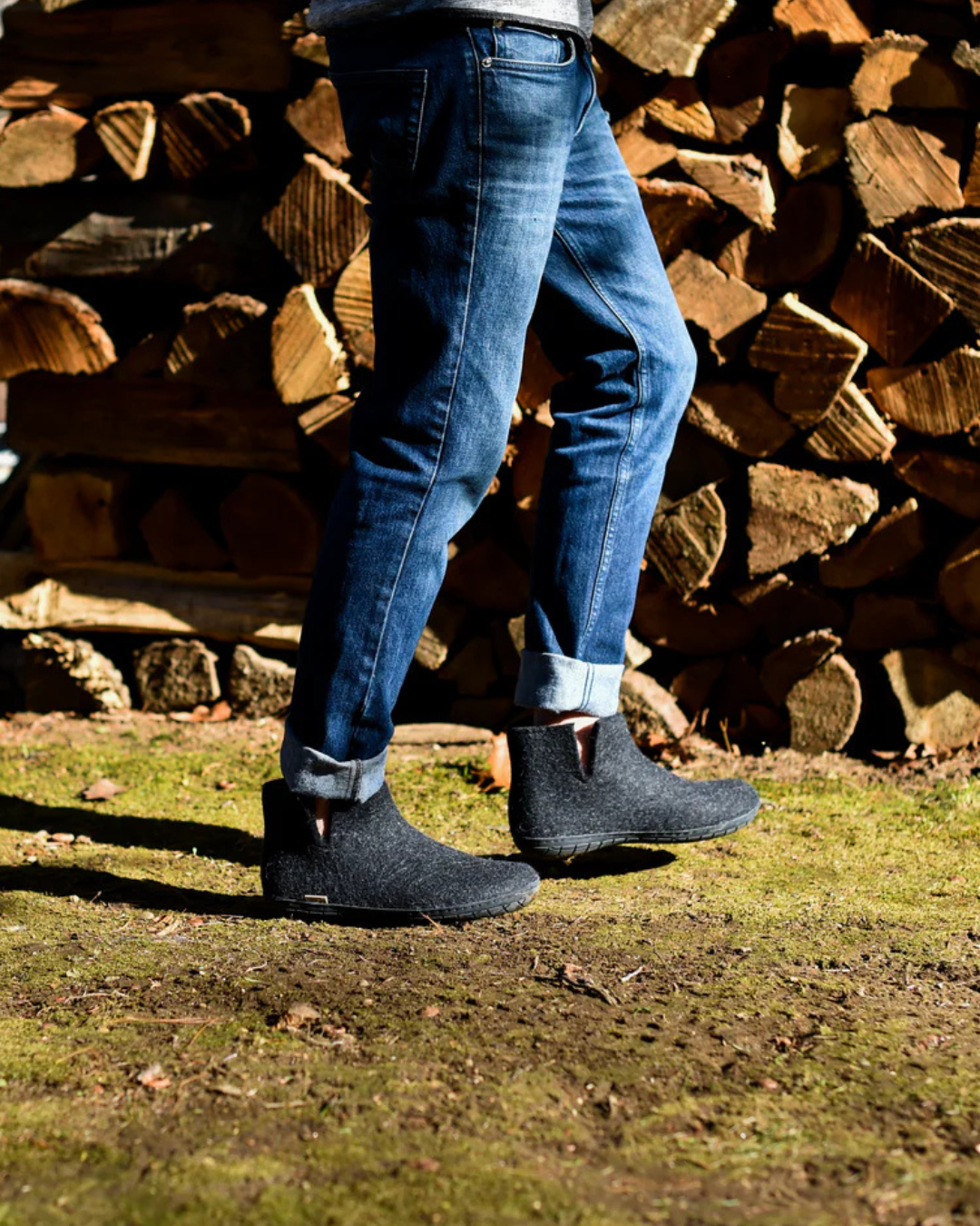 A person wearing dark gray wool felt boots and blue jeans walking outdoors on grass in front of a stacked woodpile.