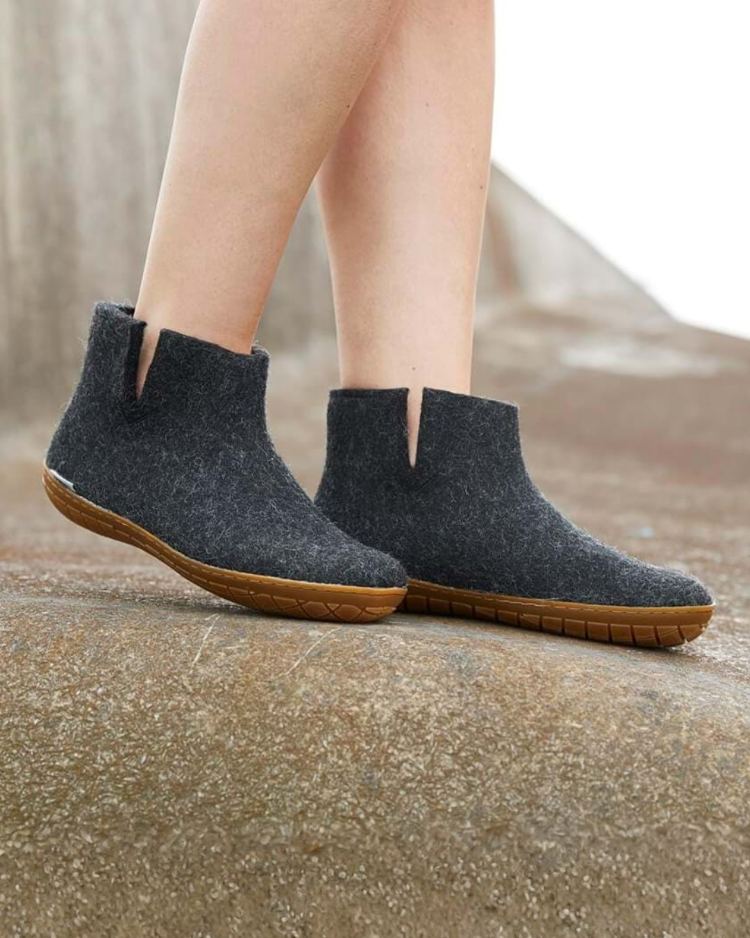 A person wearing dark gray wool felt ankle boots with brown rubber soles, standing on a textured concrete surface.