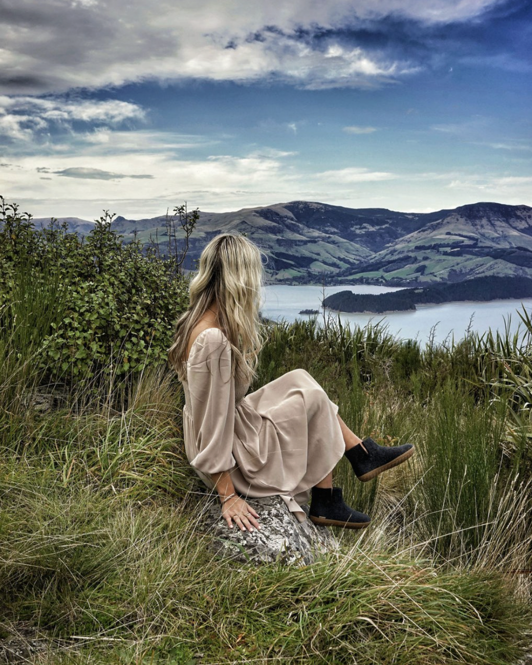 A woman with long blonde hair sits on a grassy hillside overlooking a scenic lake and mountains, wearing a loose beige dress and black wool felt ankle boots.