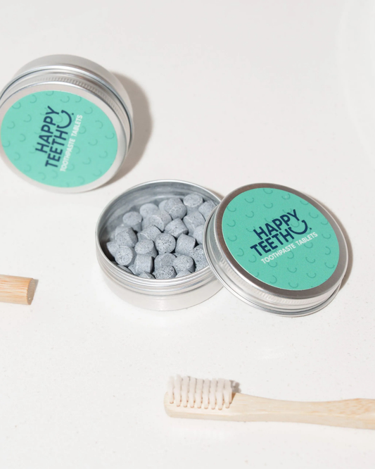 Two round silver metal tins of Happy Teeth Daily Cleanse Teeth tablets, one open showing gray tablets inside and one closed, styled on a white surface with a bamboo toothbrush and a wooden toothpick.
