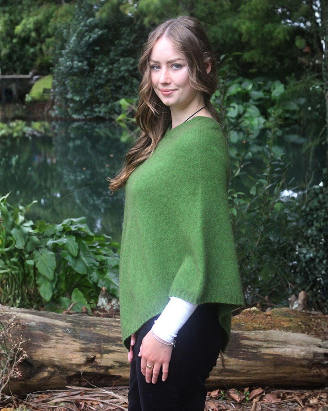 A woman wearing a possum merino green poncho over a white long-sleeve shirt and black pants, standing outdoors in front of a forest background.