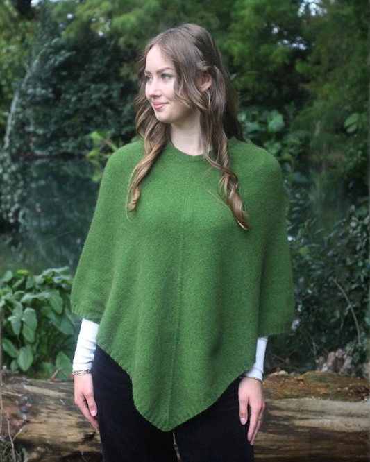 A woman wearing a possum merino green poncho over a white long-sleeve shirt and black pants, standing outdoors in front of a forest background.