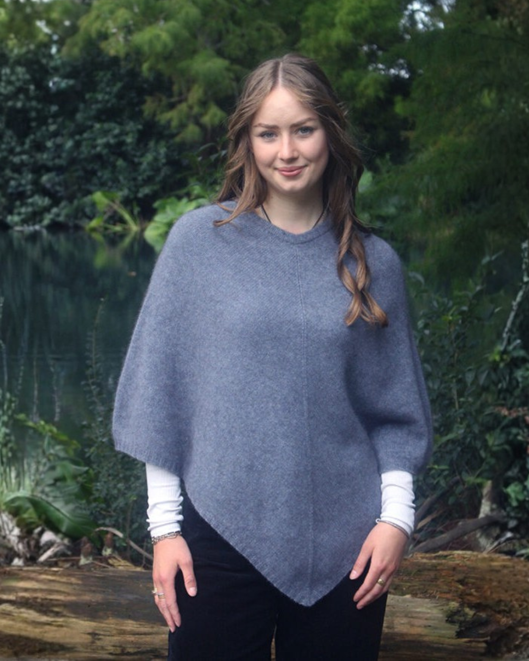 A woman wearing a slate blue-gray possum merino poncho over a white long-sleeve shirt and black pants, standing outdoors in front of a lush green forest background.