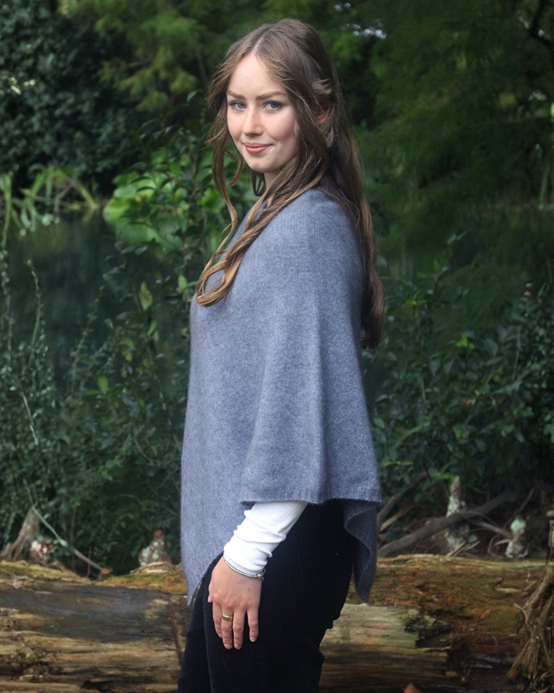 A woman wearing a light gray knit possum merino poncho over a white long-sleeve top and black pants, standing outdoors in front of a wooded background.