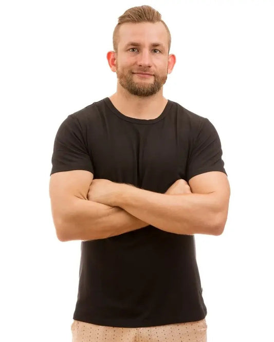 A man with short blond hair and a beard, wearing a black short-sleeve crewneck bamboo t-shirt and beige shorts, standing with his arms crossed against a solid white background.