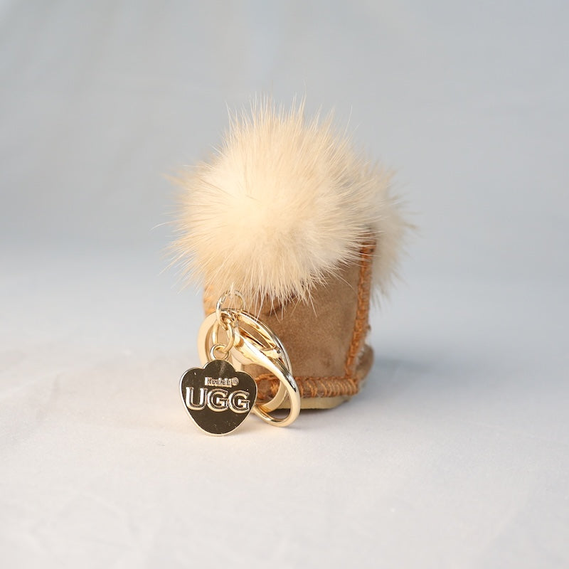 Beige UGG boot with a fluffy pom-pom and gold keychain on a light grey background