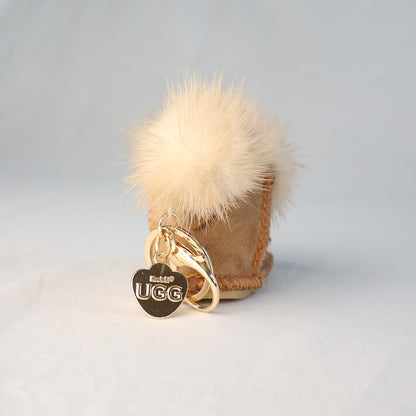 Beige UGG boot with a fluffy pom-pom and gold keychain on a light grey background