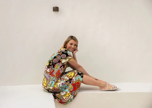 Woman wearing colourful patterned bamboo knee length dress sitting in front of white wall.