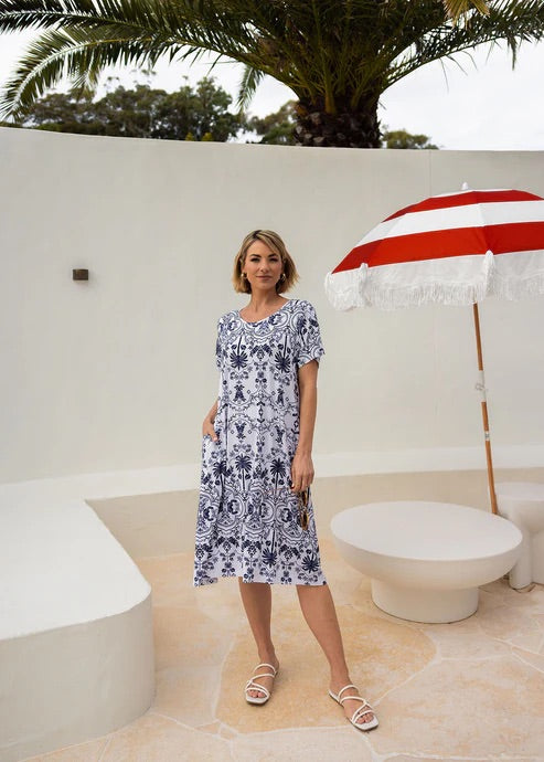Woman wearing white and blue patterned bamboo knee length dress standing near beach umbrella.