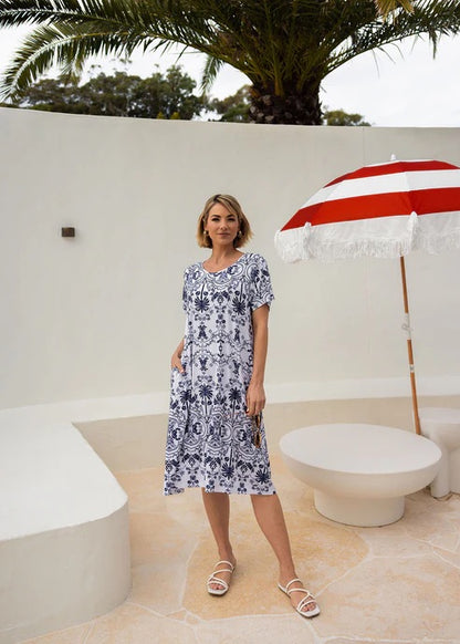 Woman wearing white and blue patterned bamboo knee length dress standing near beach umbrella.