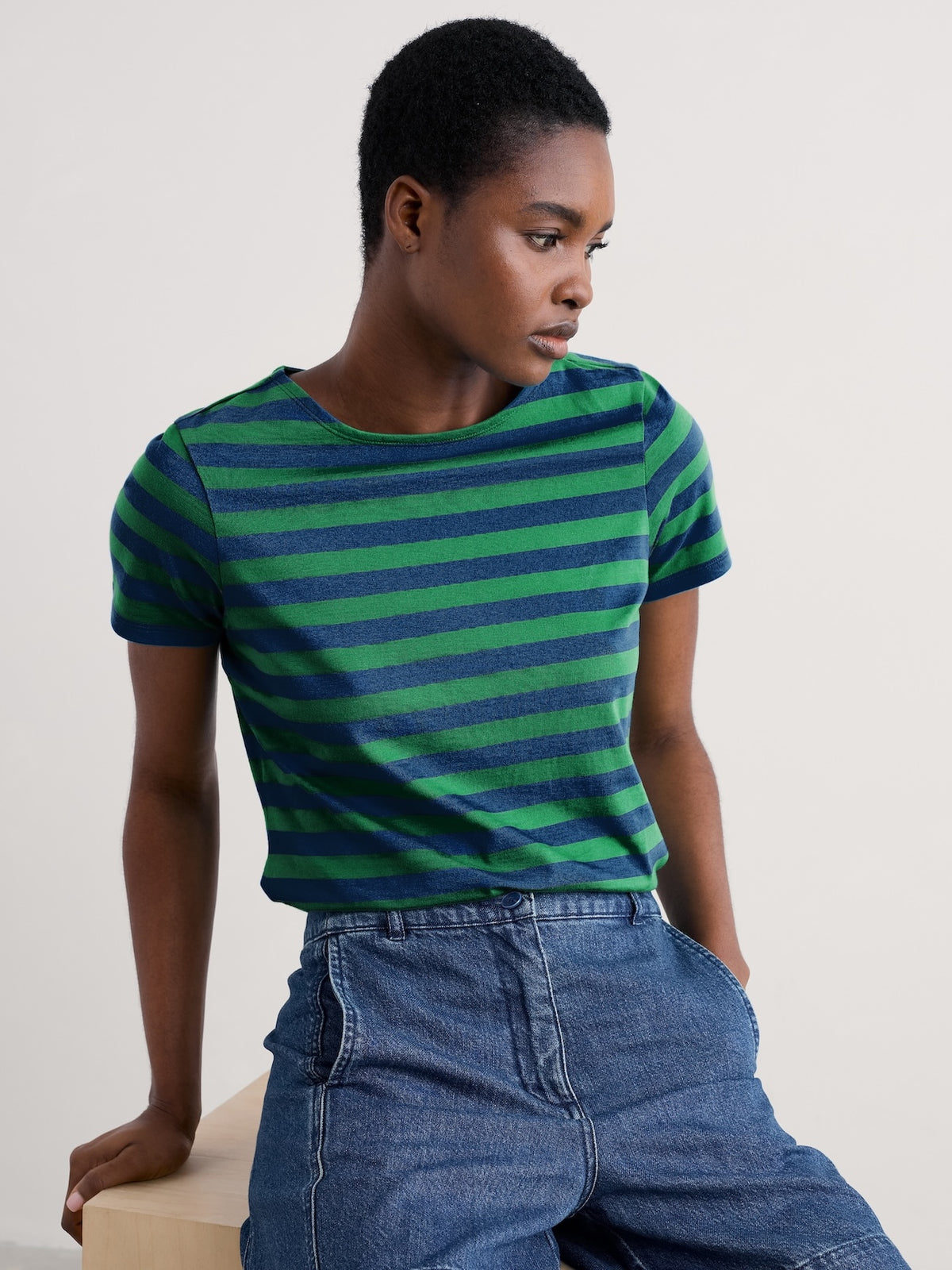 A model wearing a green and navy blue horizontally striped short-sleeve sailor t-shirt tucked into blue denim jeans, posed against a light beige studio background.
