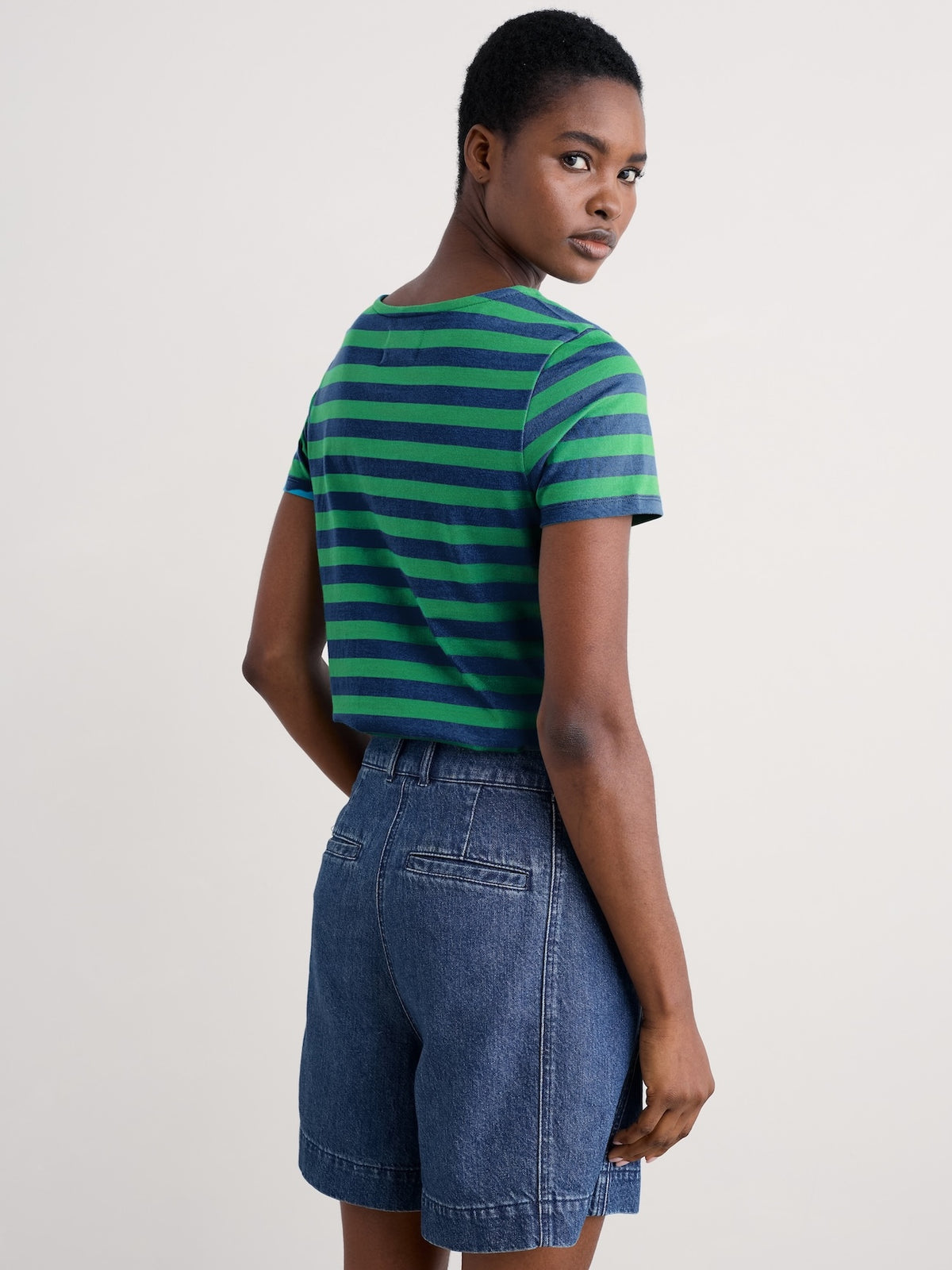 A model shown from the back wearing a green and navy blue horizontally striped short-sleeve sailor t-shirt and blue denim shorts, against a plain light gray studio background.