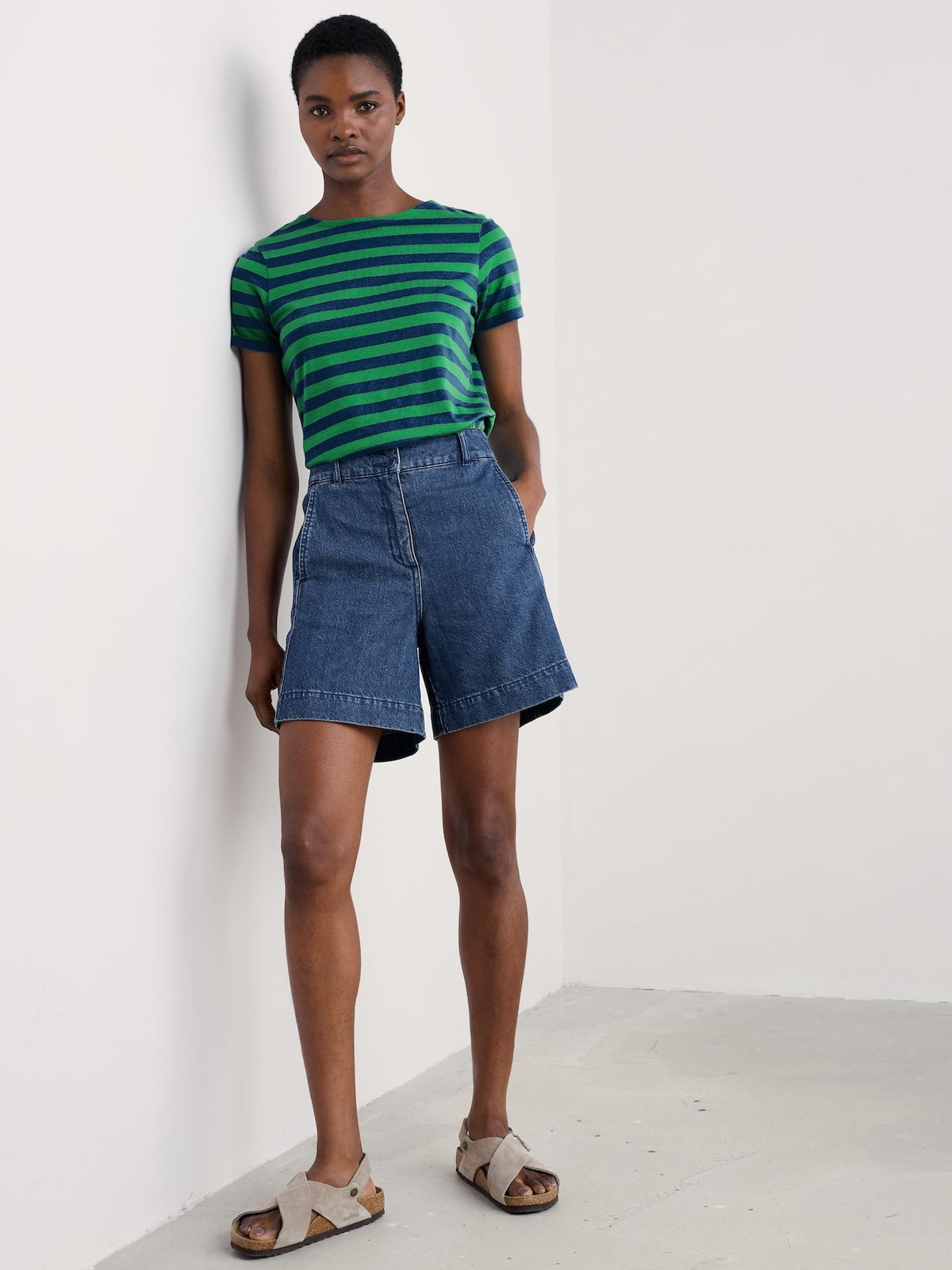 A model stands against a white wall wearing a green and black striped short-sleeve sailor t-shirt, blue denim shorts, and beige slide sandals.