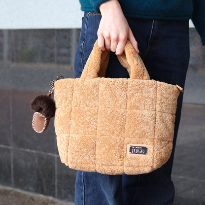 Person holding a sheepskin brown handbag with a small moccasin keychain attached, against a blurred background.