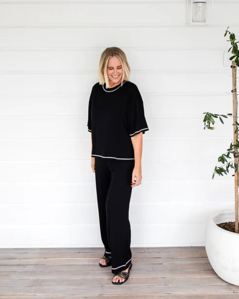 Woman wearing a black bamboo shirt and pants standing in a room with a white wall and a plant.