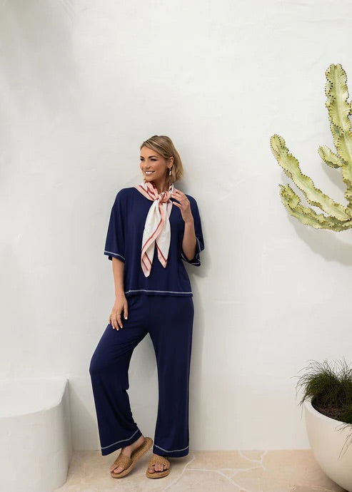 Woman in navy bamboo shirt and pants with pink scarf standing against a white wall with decorative elements.