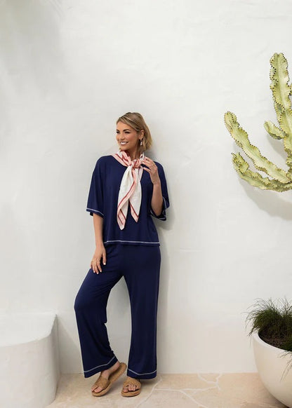 Woman in navy bamboo shirt and pants with pink scarf standing against a white wall with decorative elements.